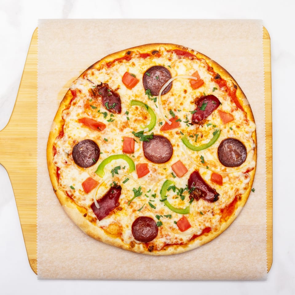 A whole pizza with pepperoni, green peppers, onions, tomatoes, and parsley on a wooden peel.