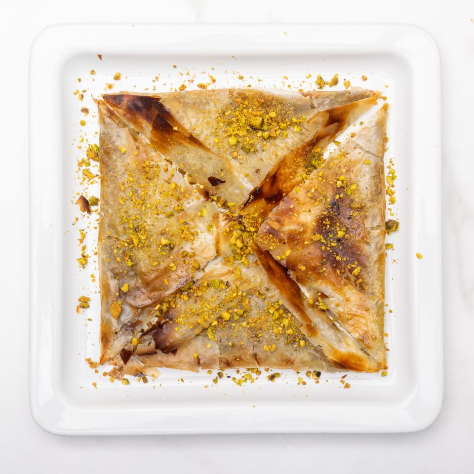 A square, golden-brown pastry, possibly baklava, sprinkled with chopped pistachios on a white plate.