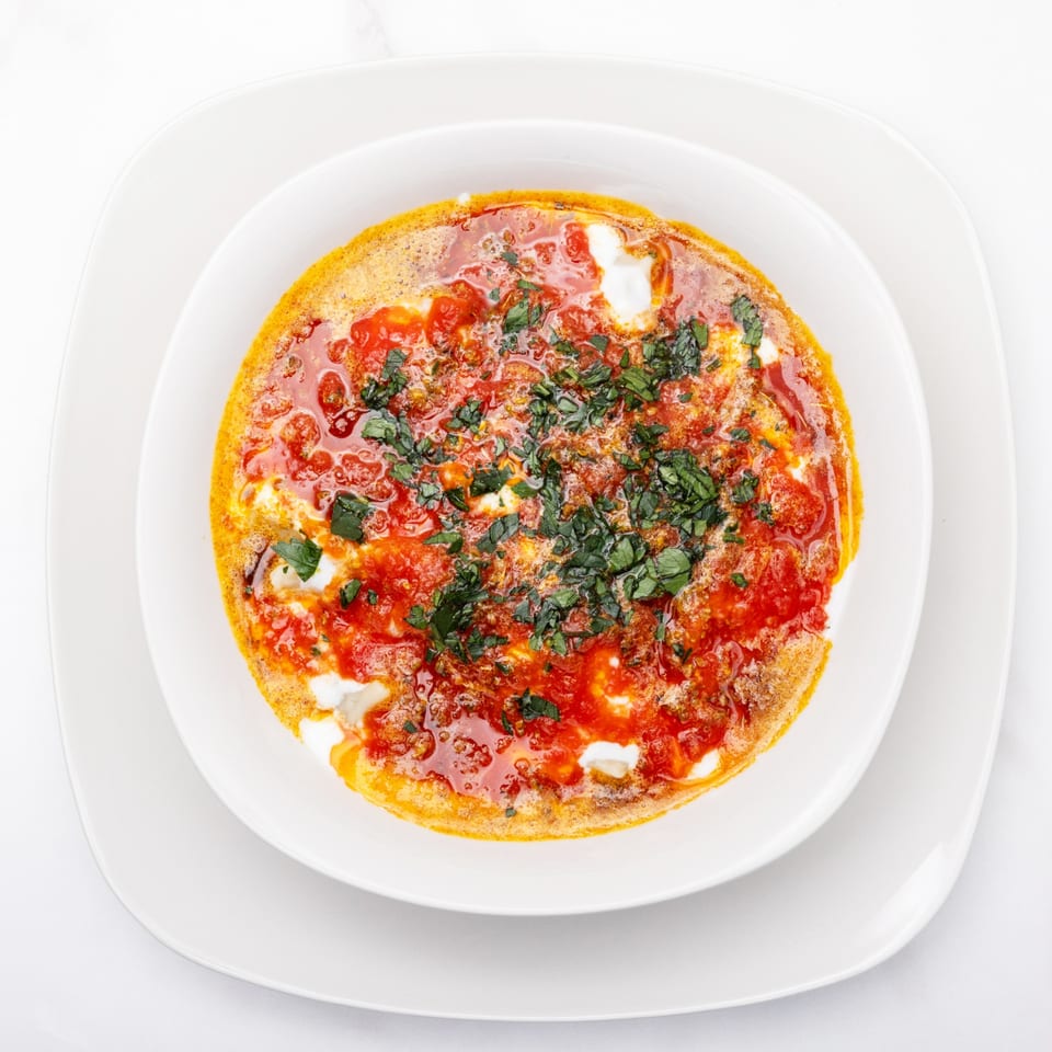 Overhead view of a shakshuka dish with red tomato sauce, white cheese, and green herbs.